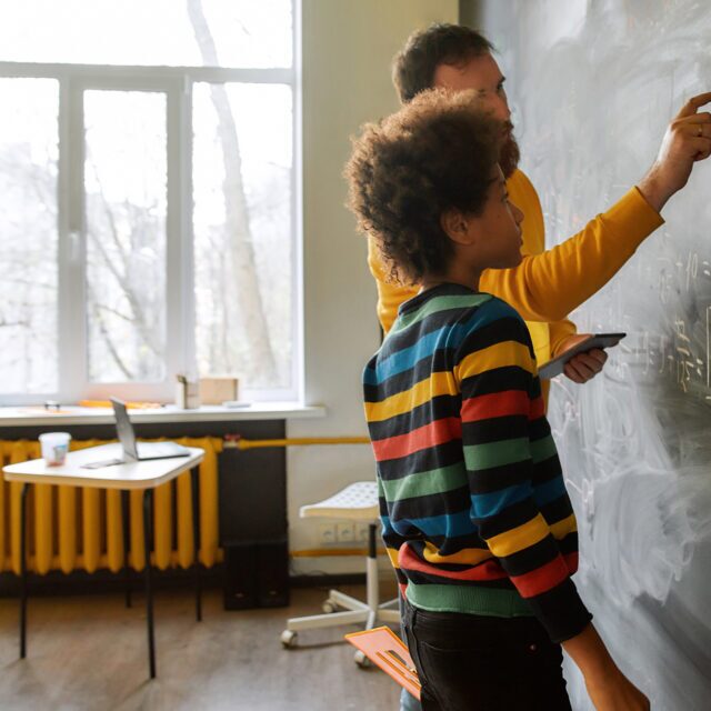student working with a teacher on math problems while standing in front of a chalk board