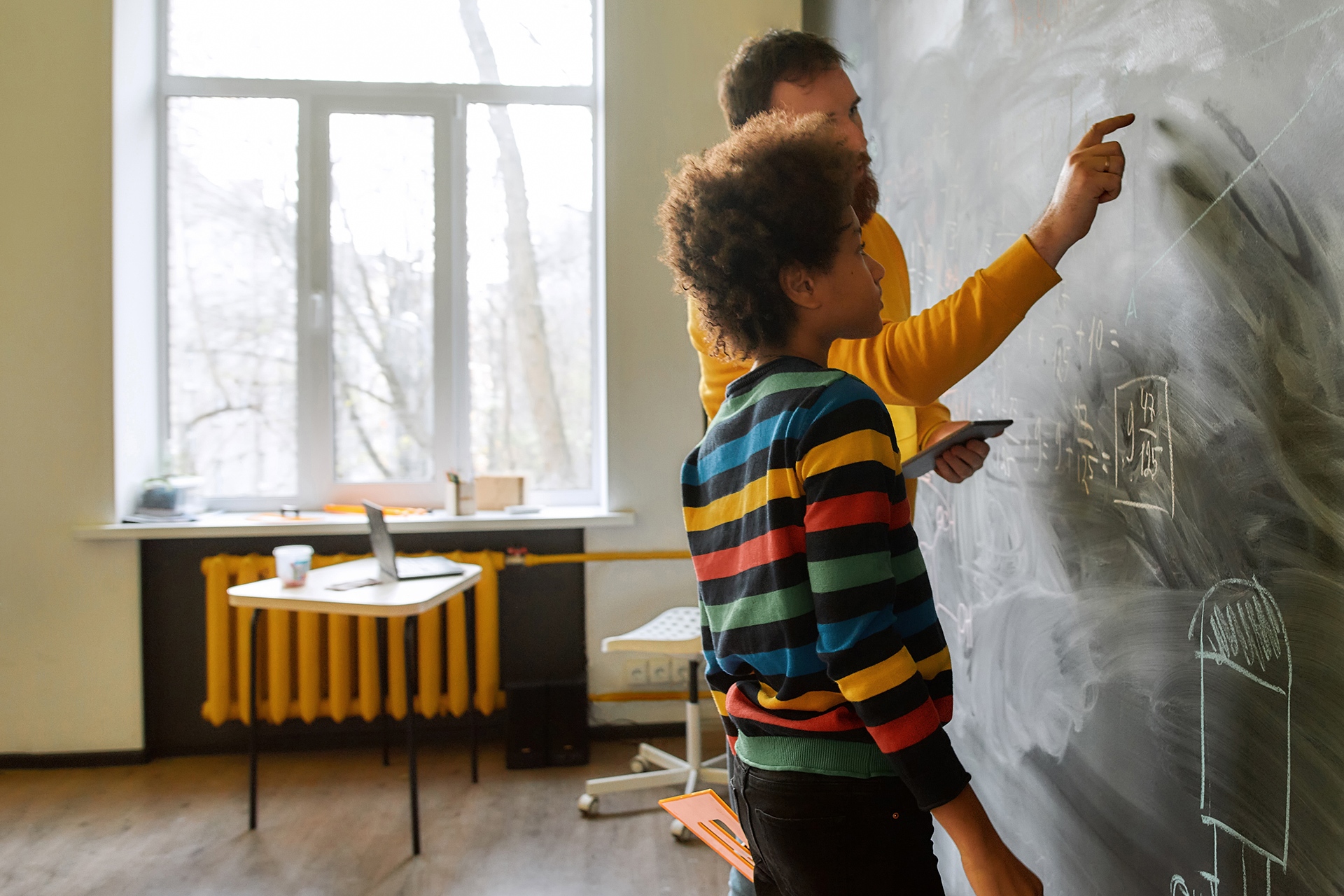student working with a teacher on math problems while standing in front of a chalk board