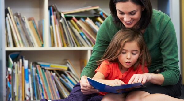 A student sounds out words while reading a book with a teacher in a classroom.