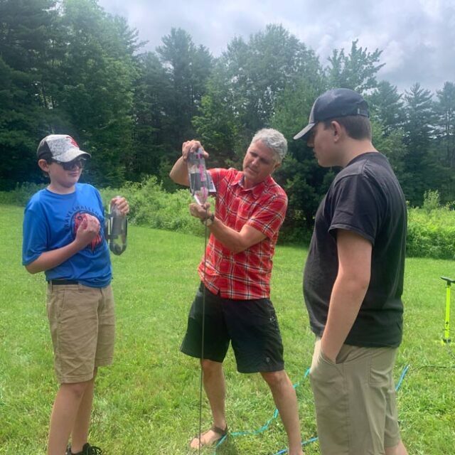 Teenage boys watch a male teacher assemble a pop bottle rocket