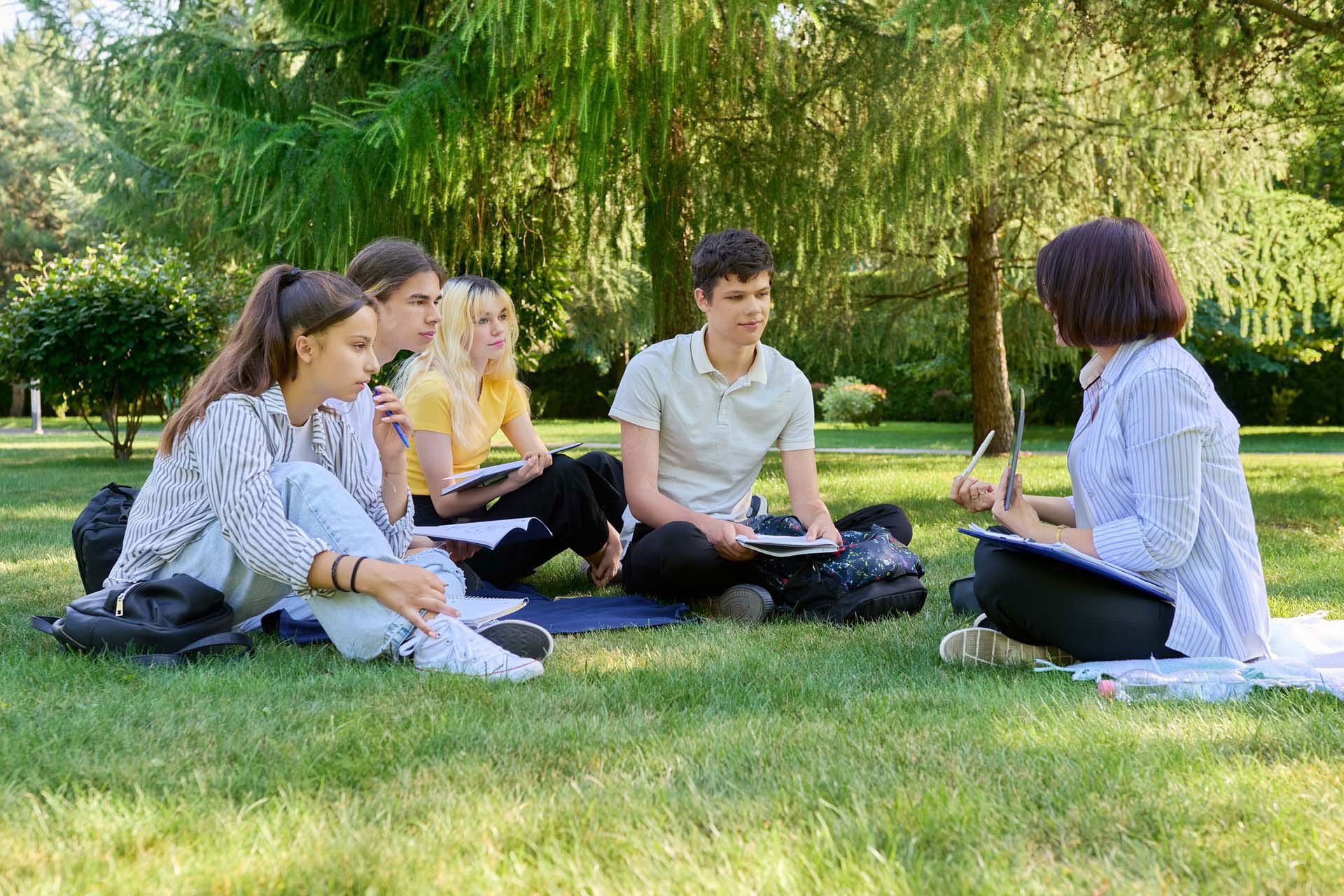 Young adults listen to a teacher while sitting outside in the grass