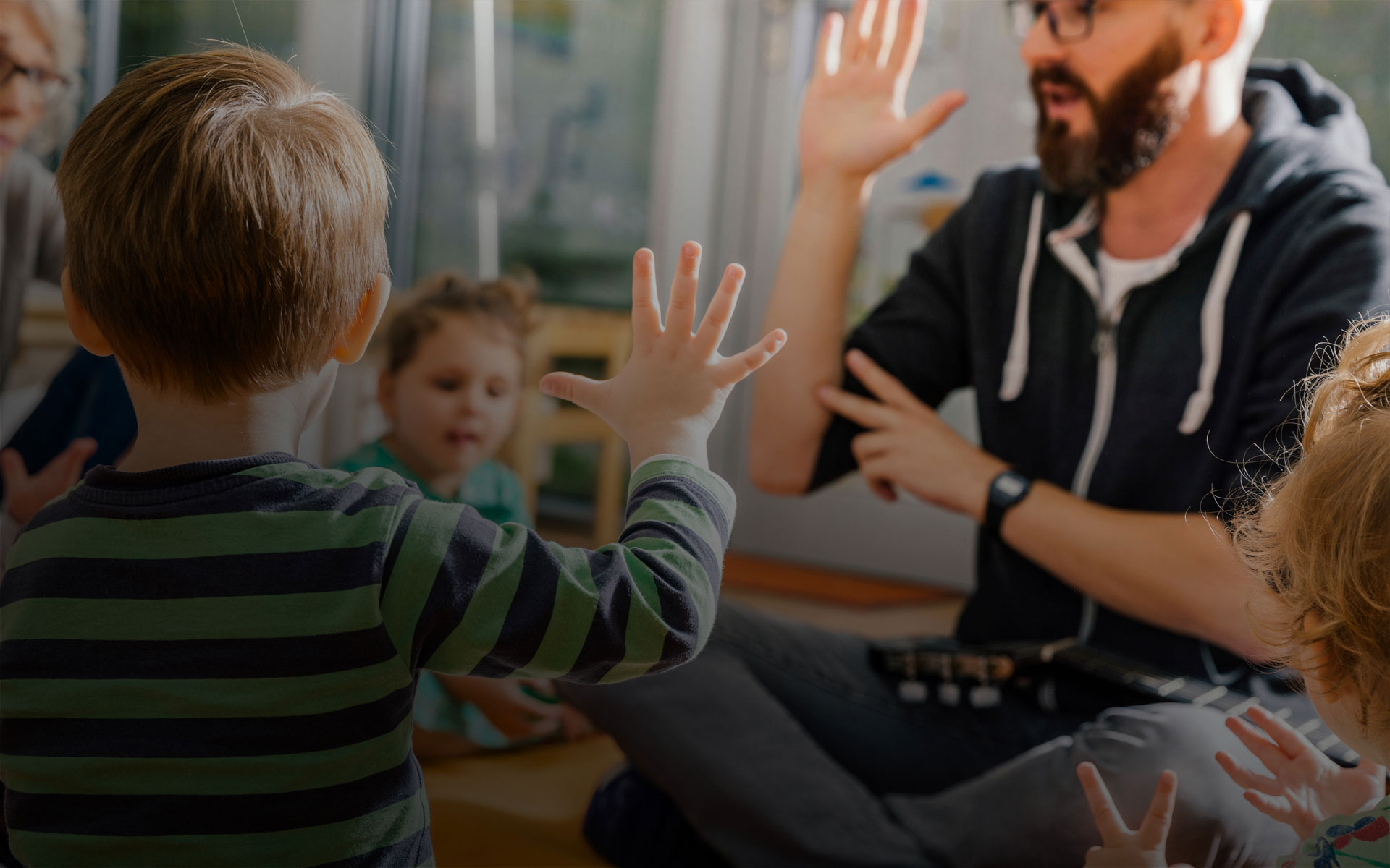 Teacher and kindergarten students sitting on the floor in a classroom, raising their hands