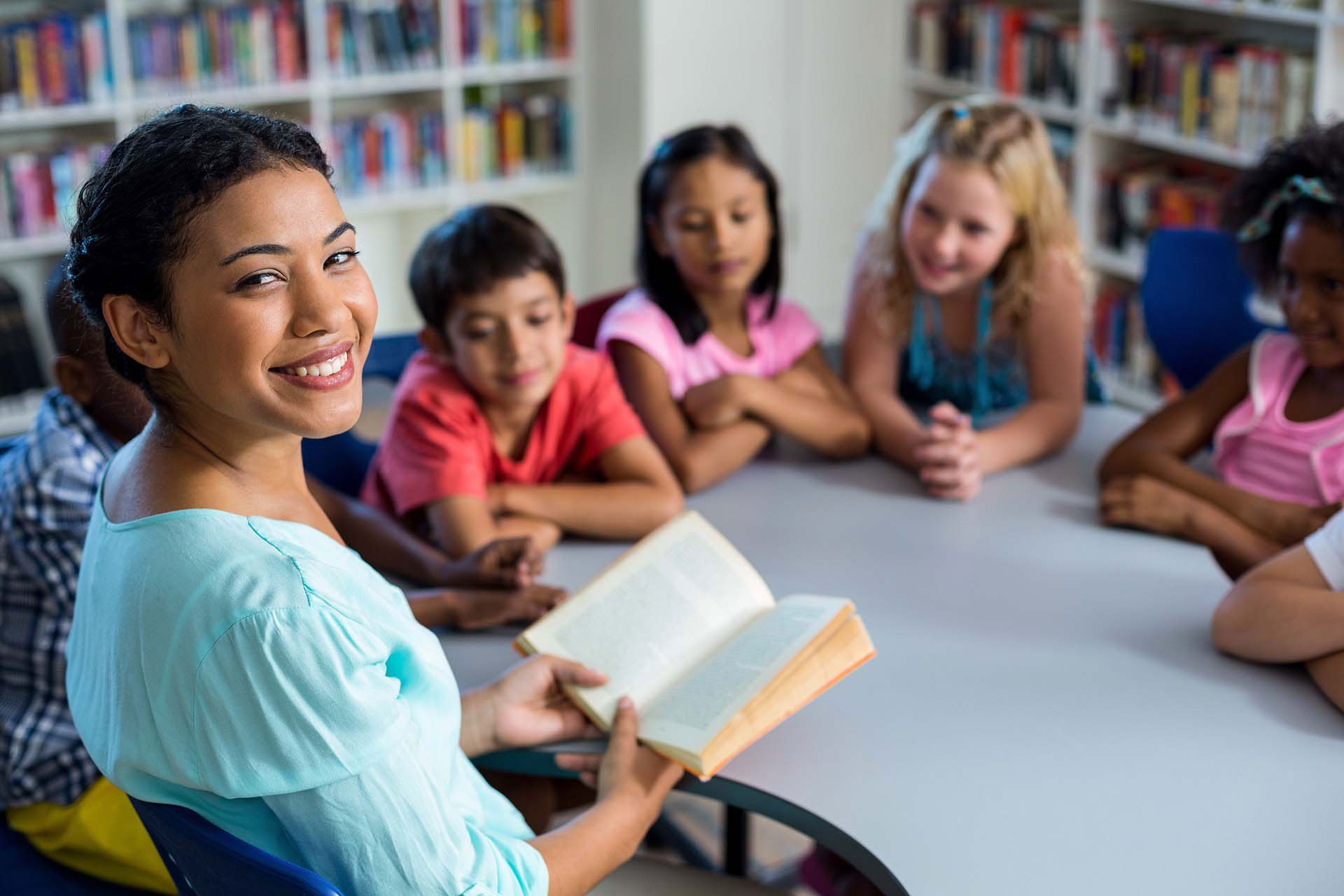 Elementary school teacher smiling and reading a book to a group of young students in a library