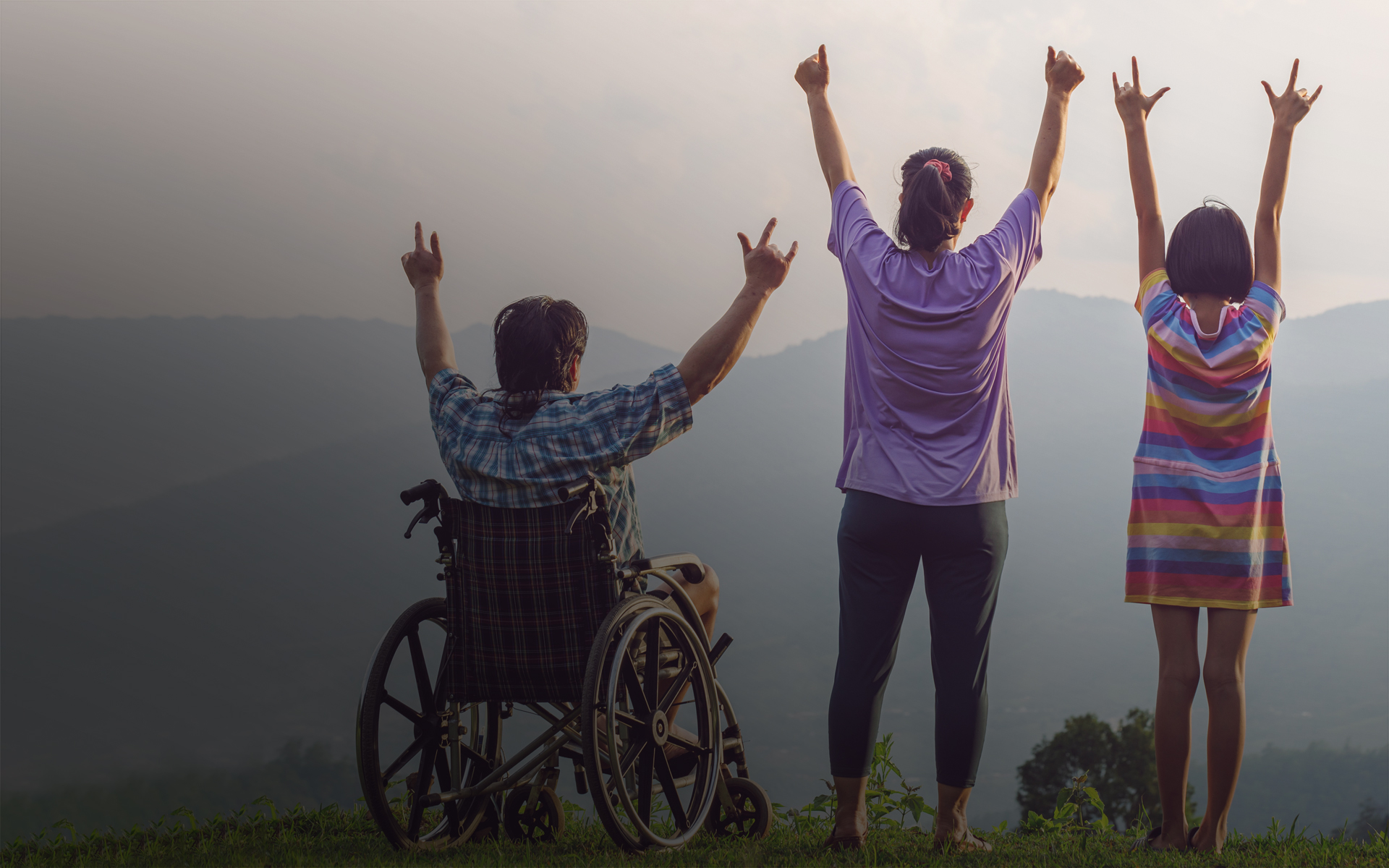 Three people, one in a wheelchair, with their arms raised and mountains in the distance