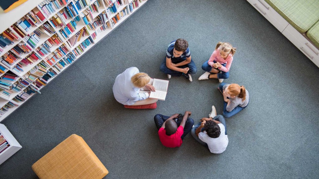 An overhead shot of a teacher reading a book to students in a library.