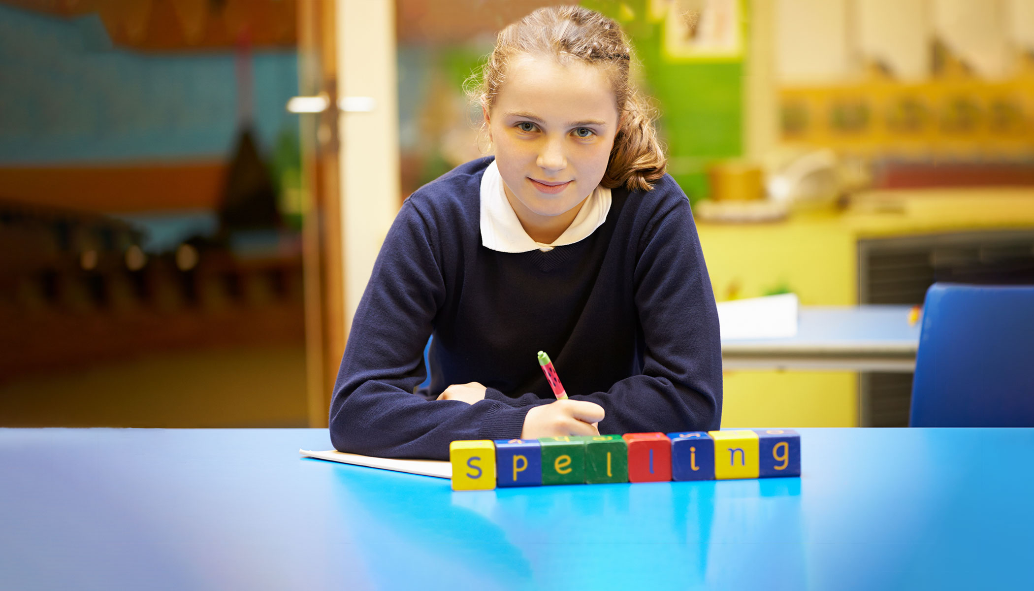 A student sits at a table in a school classroom. On the table are blocks spelling out “spelling.’